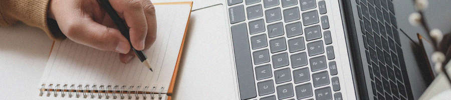 A person writing on a notebook in front of a laptop